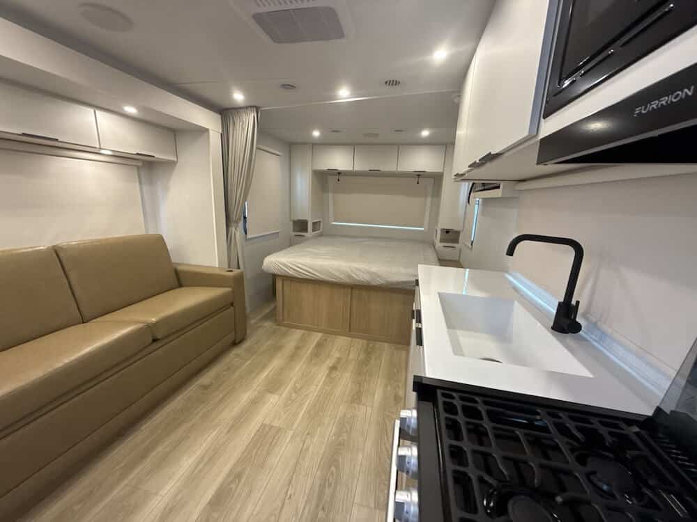 Image of the interior of the Winnebago Thrive 18FBS. The image features a sofa on the left, sink on the right and bed straight ahead. 
