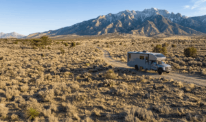 Image of a desert landscape with a Class C RV in the foreground and a mountain range behind it.